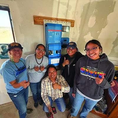 A group of students and community members smile in front of an electrical panel.